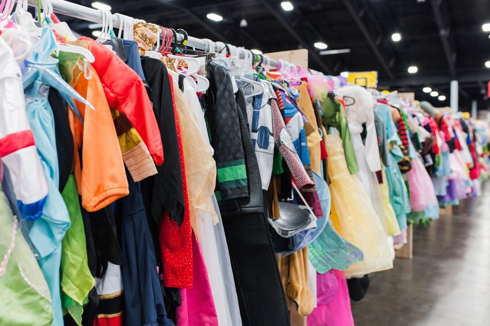 Mom wears baby and holds toddler boy clothes in each hand. Second mom shops behind her from the racks of boys clothes.